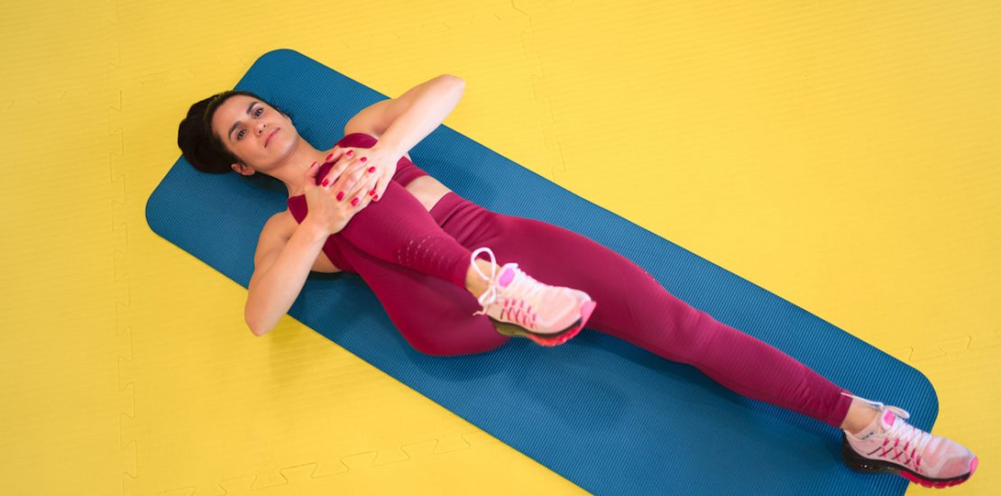 From above shot of pretty young woman in sportswear lying on stretching mat on yellow background and pulling knee to chest